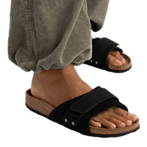 A person wearing green corduroy pants and black open-toe sandals with a cork sole and adjustable strap.