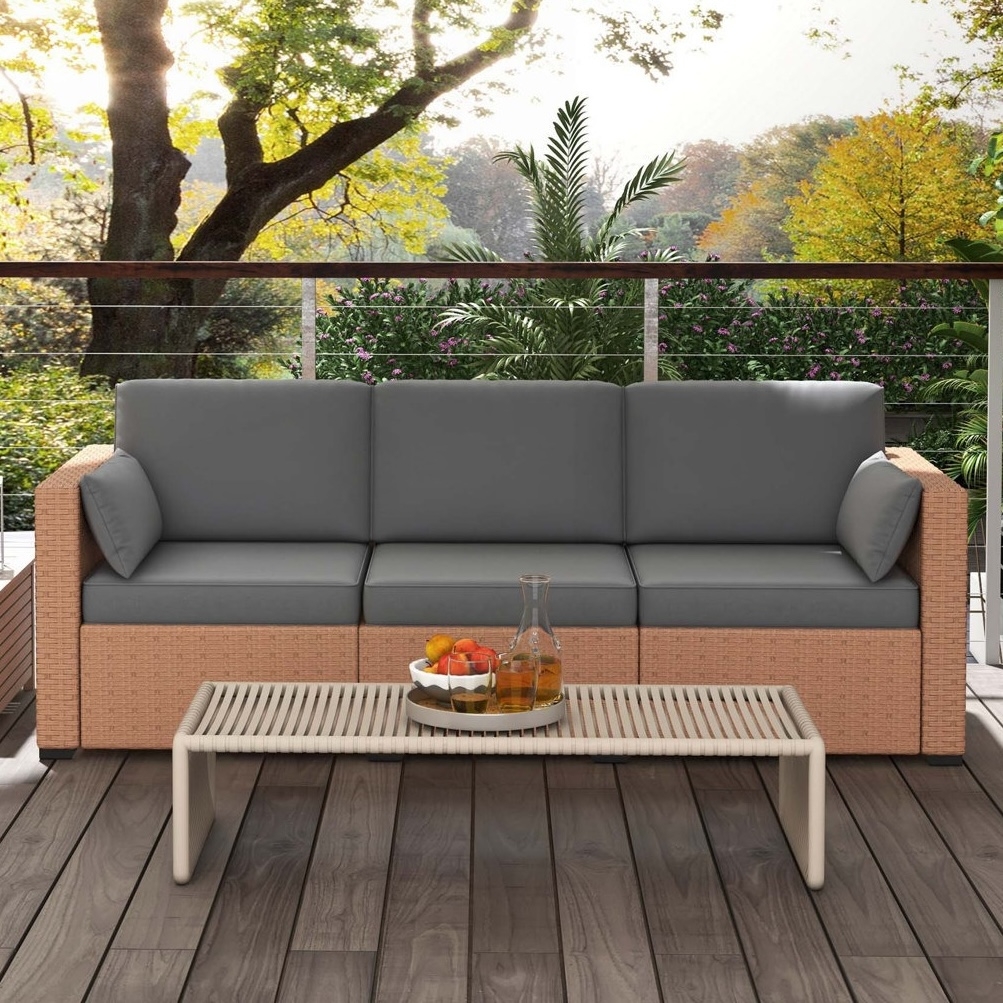 Outdoor rattan sofa with gray cushions and matching coffee table, set on a wooden deck with lush greenery and trees in the background.