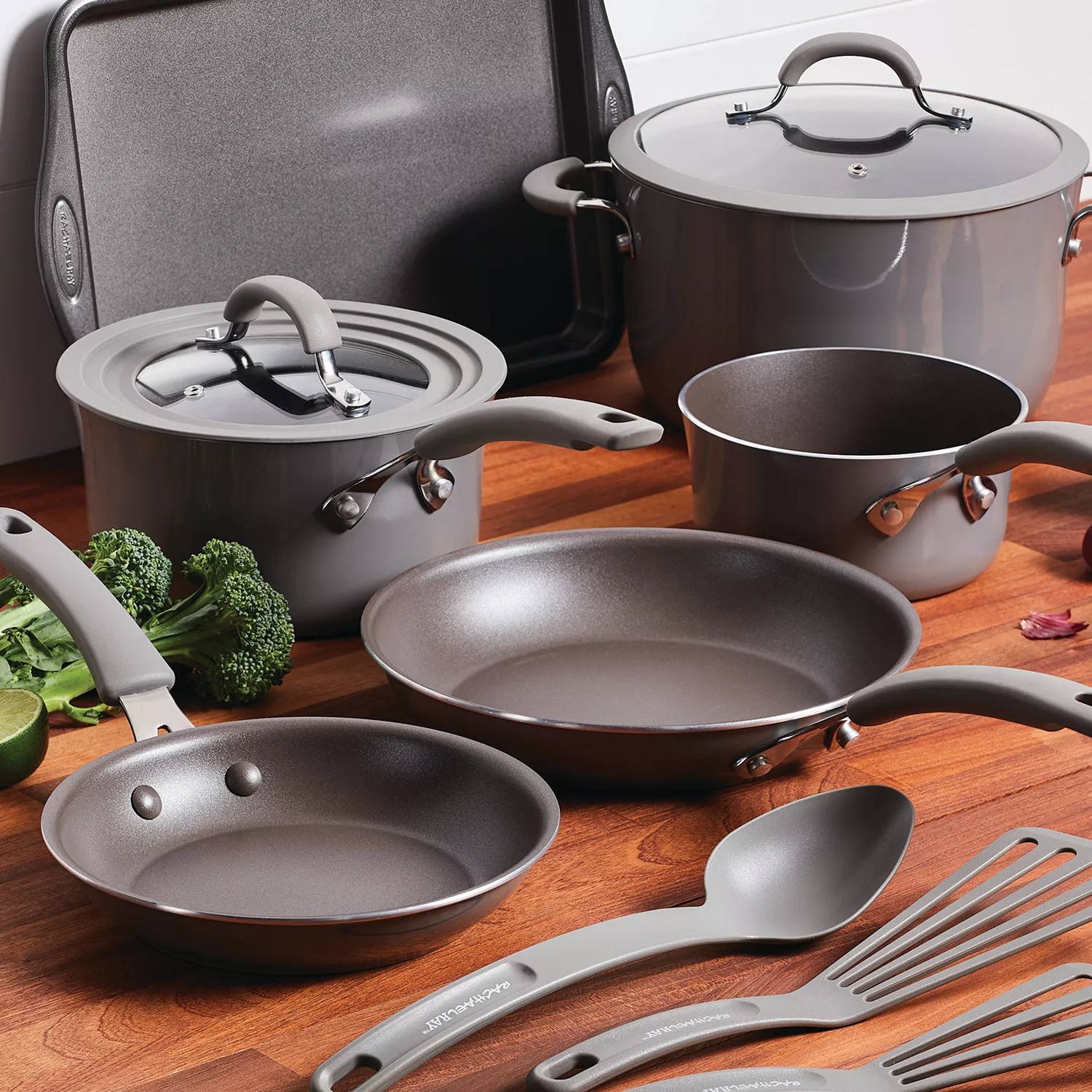 Cookware set including pots, pans, and cooking utensils with gray non-stick surfaces and matching lids, displayed on a wooden countertop with vegetables nearby.