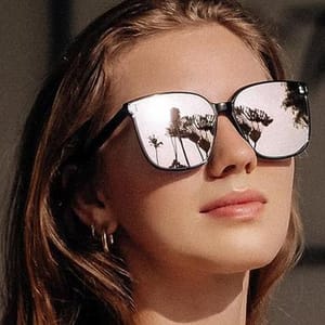 A person is wearing large, reflective sunglasses with palm tree reflections visible on the lenses, paired with small hoop earrings.