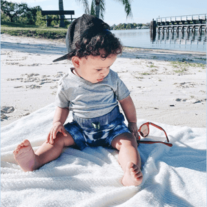 A young child sits on a beach blanket wearing a gray shirt and blue shorts, with a black cap worn backward. Nearby are a pair of sunglasses, and the background features palm trees, sand, and water.