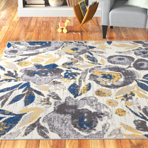 8' x 10' floral area rug with a gray, blue, and yellow pattern on a neutral background, placed on wooden flooring beside a gray chair and a magazine holder in a cozy room setting.