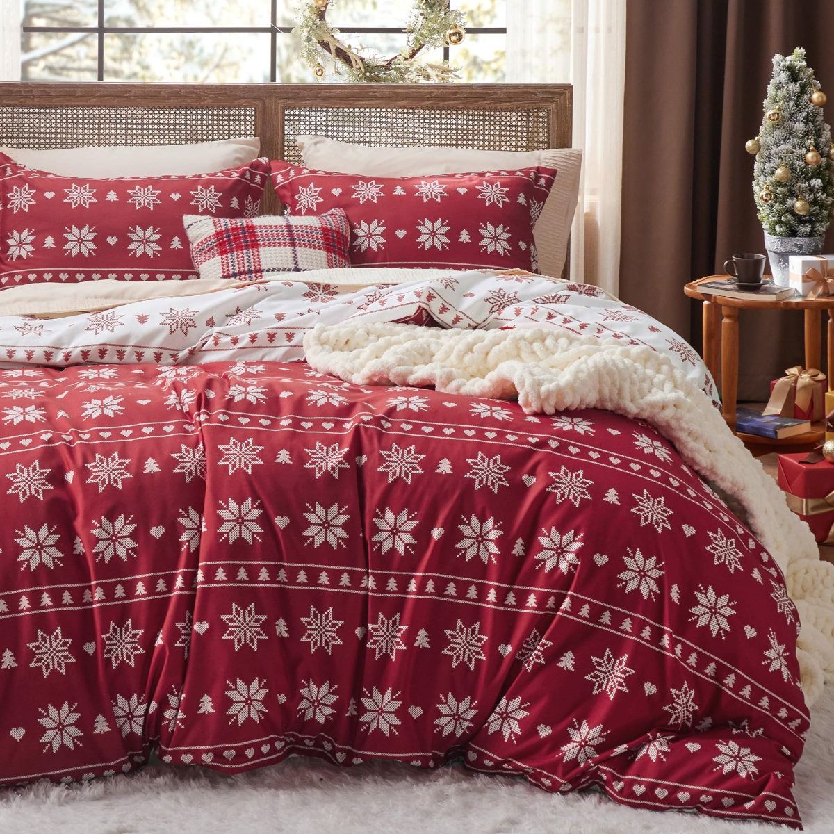 Holiday-themed bedding set with red and white snowflake patterns, accompanied by a white knit blanket. Decor includes a small decorated Christmas tree and wrapped gifts.