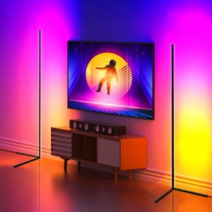 Two slim, vertical LED floor lamps emit adjustable colored lighting in two corners of a room, enhancing the ambiance around a mounted television.
