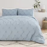 A light blue, textured comforter set with a tufted design is displayed on a bed, accompanied by matching pillow shams. The background features neutral decor, including a plant, vases, and a wicker basket.