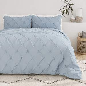 A light blue, textured comforter set with a tufted design is displayed on a bed, accompanied by matching pillow shams. The background features neutral decor, including a plant, vases, and a wicker basket.