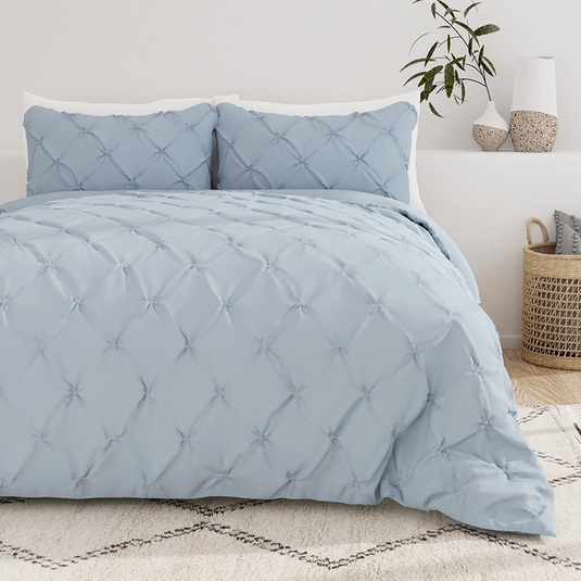 A light blue, textured comforter set with a tufted design is displayed on a bed, accompanied by matching pillow shams. The background features neutral decor, including a plant, vases, and a wicker basket.