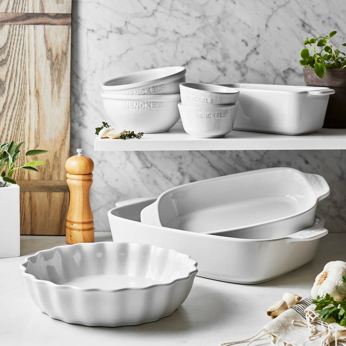 Set of white ceramic bakeware including bowls, dishes, a pie dish, and a pepper grinder, displayed on a marble and wood kitchen counter with herbs.