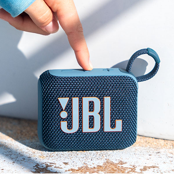 A compact JBL Bluetooth speaker with a blue woven texture and a loop for carrying, shown being pointed at by a finger.