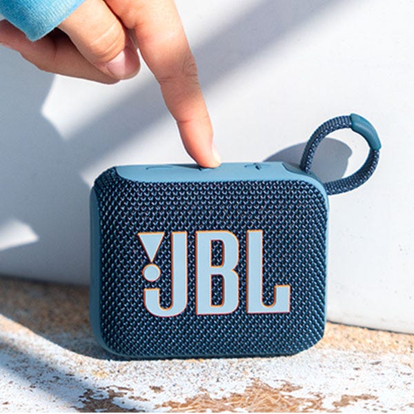 A compact JBL Bluetooth speaker with a blue woven texture and a loop for carrying, shown being pointed at by a finger.