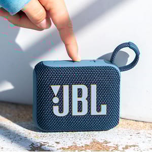 A compact JBL Bluetooth speaker with a blue woven texture and a loop for carrying, shown being pointed at by a finger.