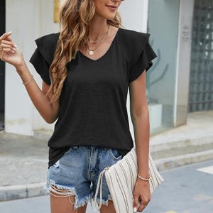 A woman is wearing a black V-neck T-shirt paired with denim shorts and a white clutch. She has layered necklaces and bangles.