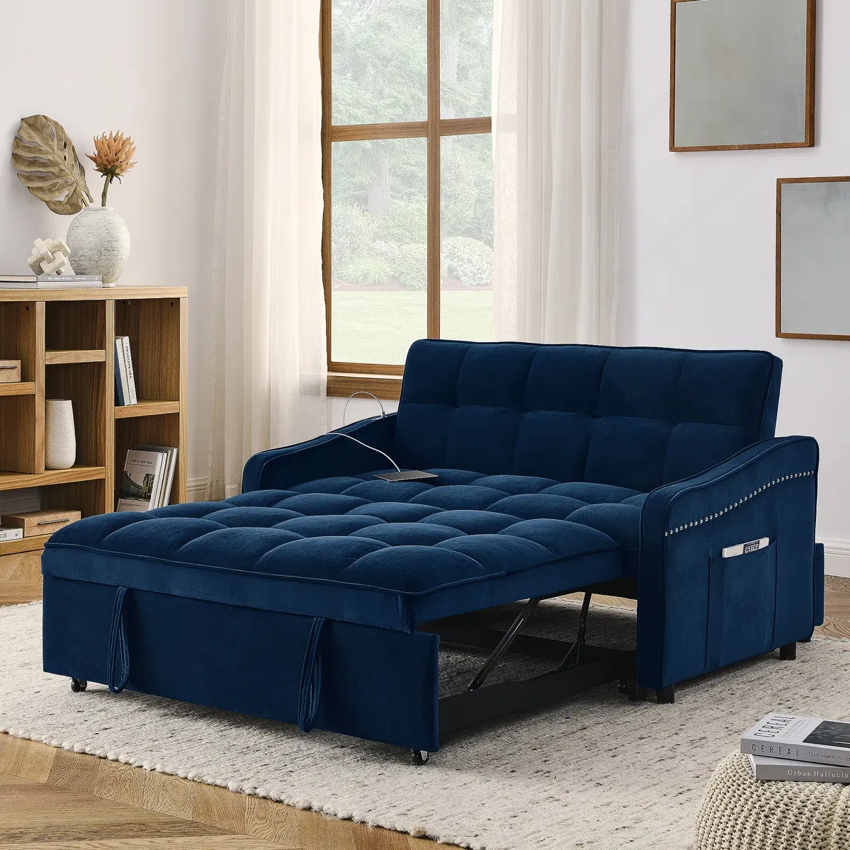A dark blue upholstered sleeper sofa is extended into a bed, featuring tufted cushions and nailhead trim. It's set in a living room with a window, bookshelf, and light decor.