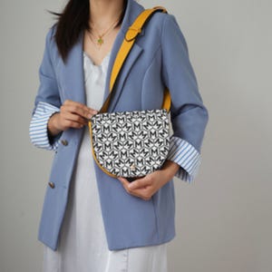 A person is wearing a blue striped blazer and holding a black and white MKF belt bag with a prominent monogram pattern and a contrasting yellow strap.