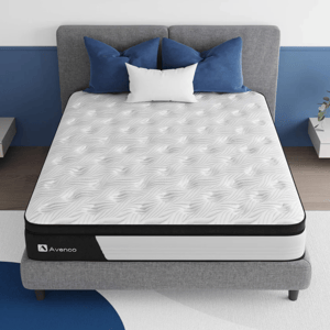 A mattress with a textured white surface and black border, branded \“Avenco,\“ is placed on a gray upholstered bed. Blue and white pillows complement the setup in a modern bedroom décor.