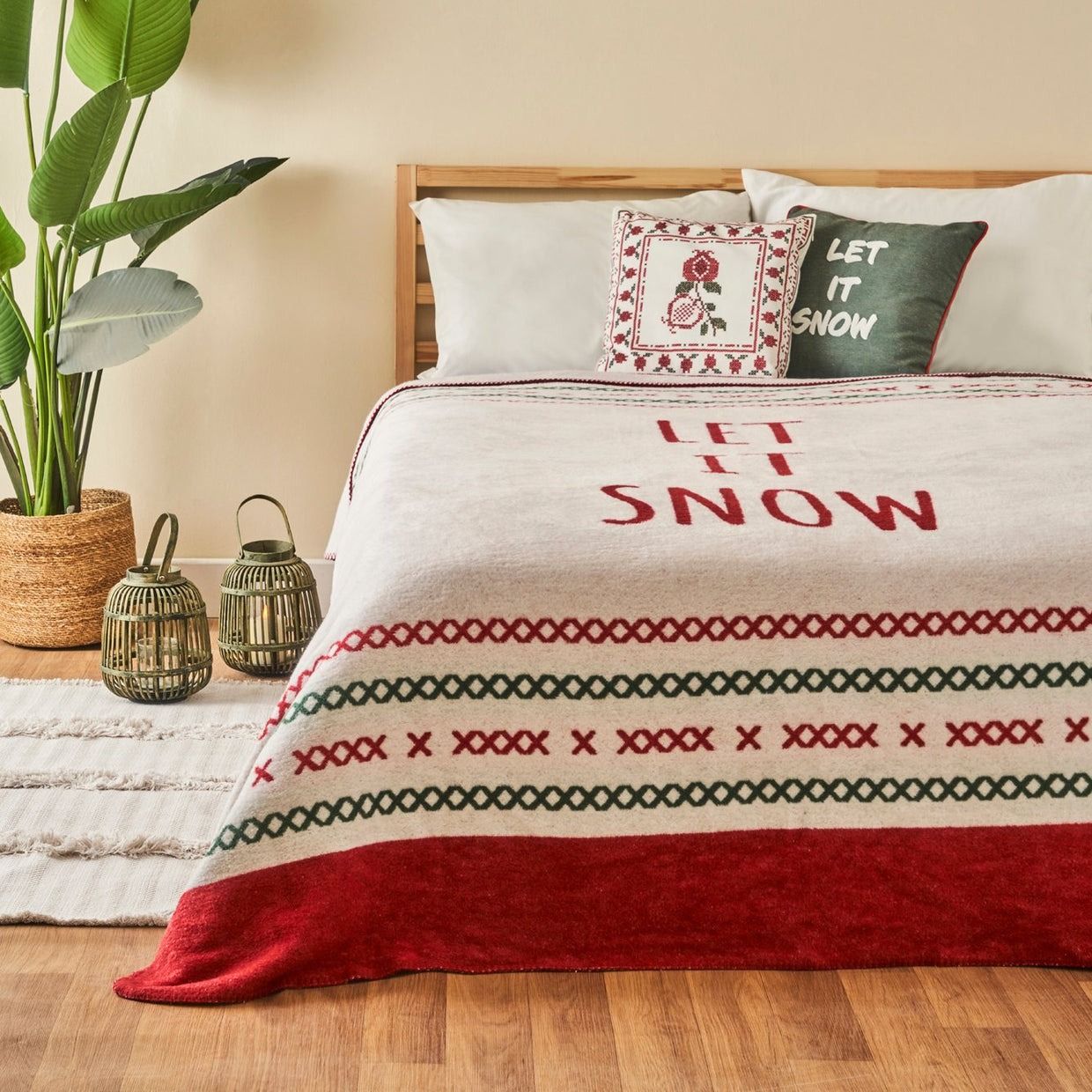 A cozy bedroom features a \“LET IT SNOW\“ themed blanket and pillows with festive designs. The room is accented with decorative lanterns, a potted plant, and a textured rug, creating a warm, inviting atmosphere.