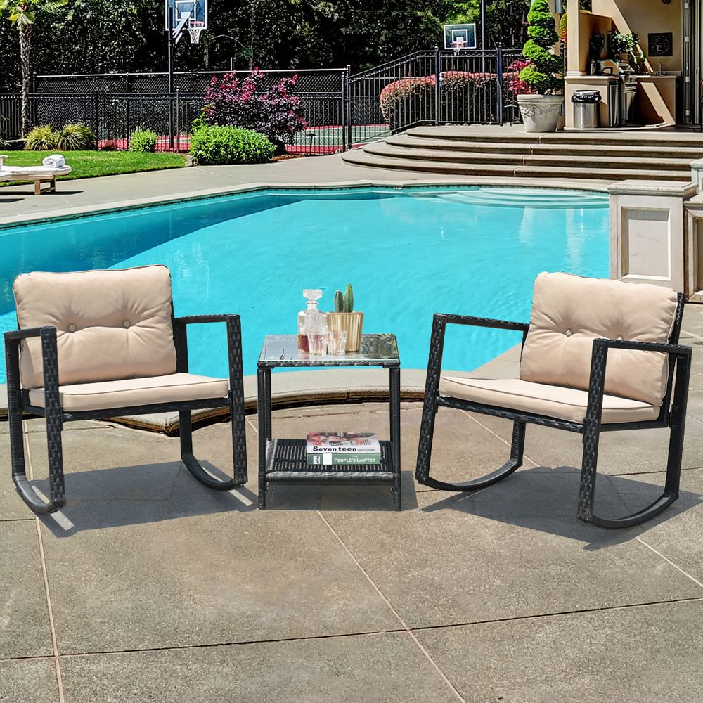 Outdoor seating set with two beige-cushioned rocking chairs and a glass-top side table. The setup is placed on a poolside patio with additional items like a decanter, glasses, and magazines on the table. In the background, there's a pool and basketball court.