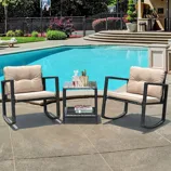Outdoor seating set with two beige-cushioned rocking chairs and a glass-top side table. The setup is placed on a poolside patio with additional items like a decanter, glasses, and magazines on the table. In the background, there's a pool and basketball court.