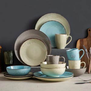 A collection of ceramic dinnerware including plates, bowls, and mugs in shades of beige, blue, and gray with a speckled design is displayed on a tabletop.