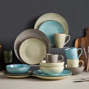 A collection of ceramic dinnerware including plates, bowls, and mugs in shades of beige, blue, and gray with a speckled design is displayed on a tabletop.