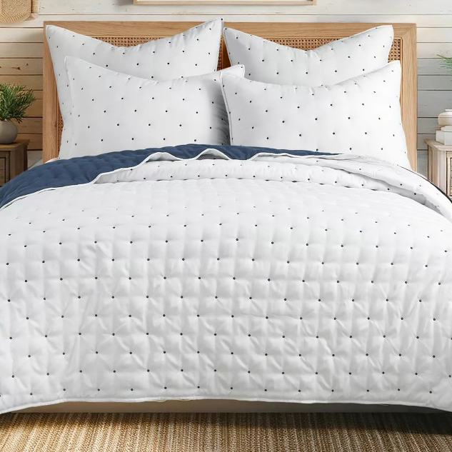 A neatly made bed features a white quilted comforter and matching pillows, all dotted with small black patterns. The bedding includes a dark blue blanket folded at the edge.