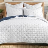 A neatly made bed features a white quilted comforter and matching pillows, all dotted with small black patterns. The bedding includes a dark blue blanket folded at the edge.