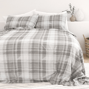 A bed with a gray plaid-patterned duvet cover and matching pillowcases, alongside white pillows. The setup includes a minimalist decor with vases and a potted plant on a bedside table.