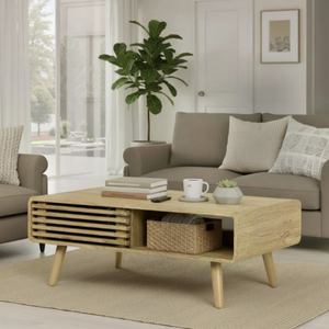 A modern living room features a wooden coffee table with a slatted design, accompanied by a gray sofa with assorted pillows. The table includes a wicker basket and books, placed on a beige area rug, with a potted plant and window in the background.