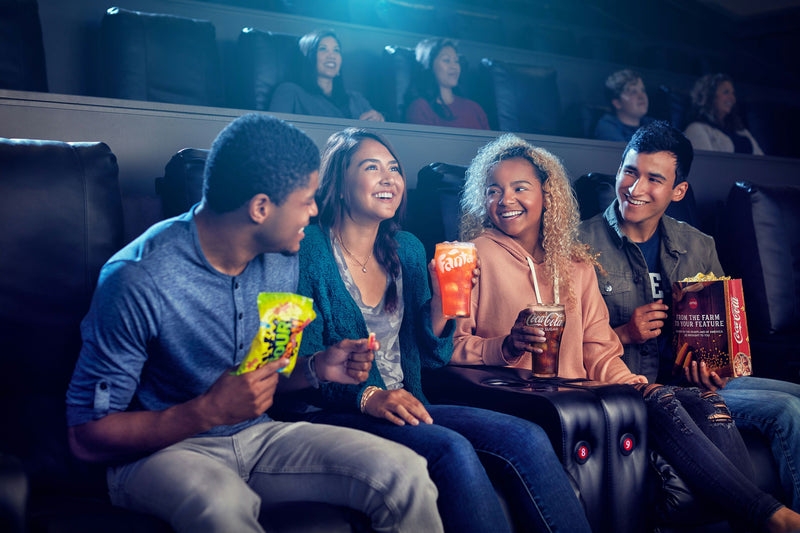 Four friends are enjoying a movie in a theater, holding soft drinks and popcorn.