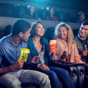 Four friends are enjoying a movie in a theater, holding soft drinks and popcorn.