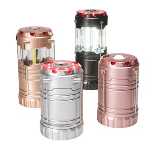 Four pop-up lanterns are shown in different metallic colors: gold, black, silver, and rose gold. The black lantern is expanded, emitting LED light, while the others are closed. Each has a top handle for carrying or hanging.