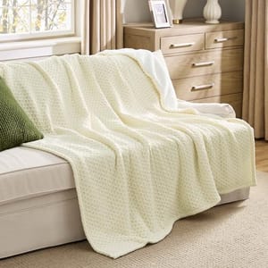 A 60\“ dual-textured cream-colored throw blanket drapes over a beige sofa with a textured pattern. It contrasts with a green cushion and is set in a cozy living room with wooden furniture and neutral curtains.