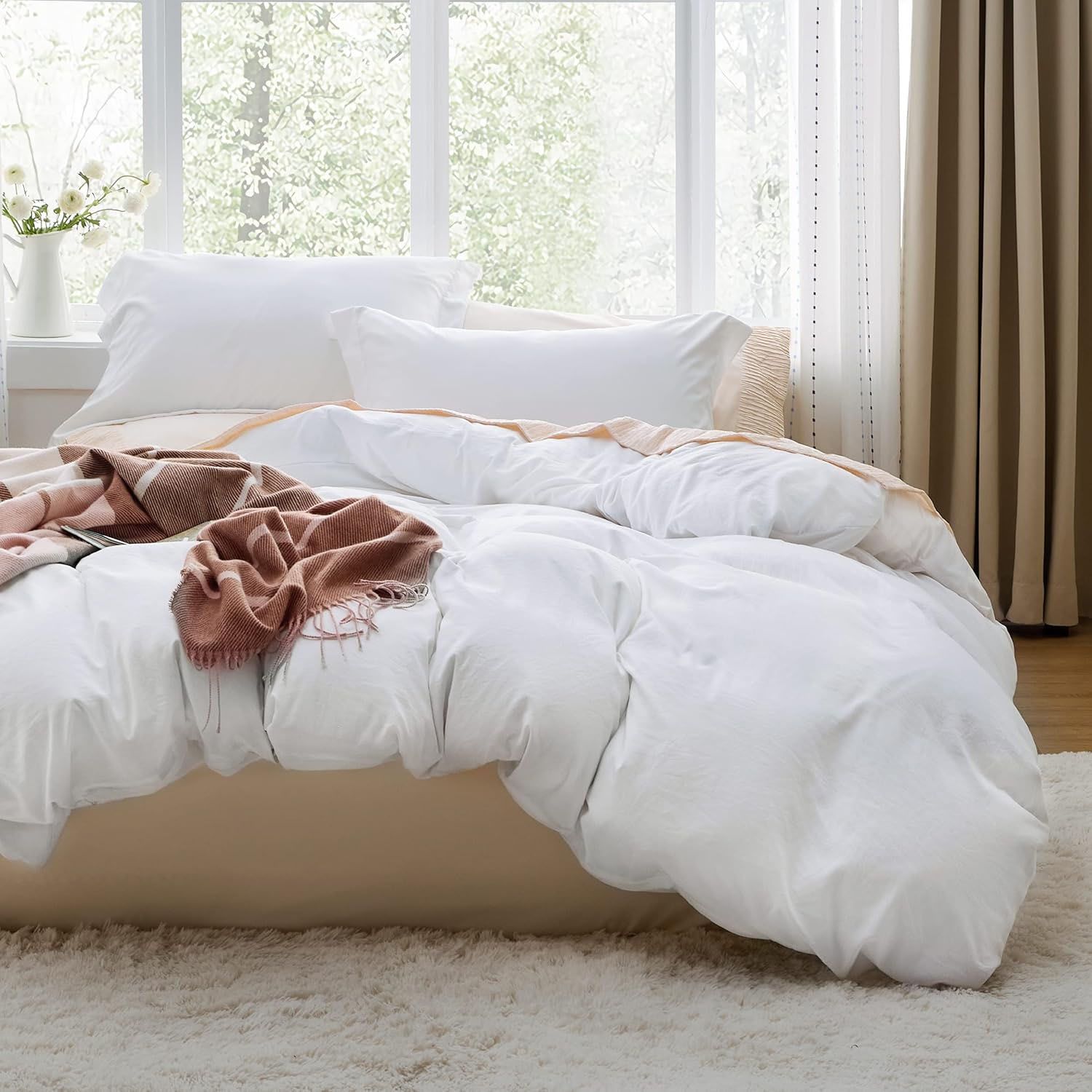 A cozy bedroom setup featuring a white comforter and pillows on a bed, accented with a brown blanket. There is a window in the background with curtains pulled to the side, and a vase with flowers on the windowsill.