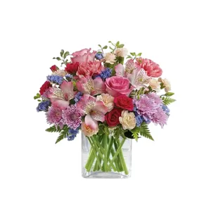 A vibrant flower arrangement featuring pink roses, red roses, pink alstroemeria, pink chrysanthemums, carnations, blue accents, and greenery in a square glass vase.