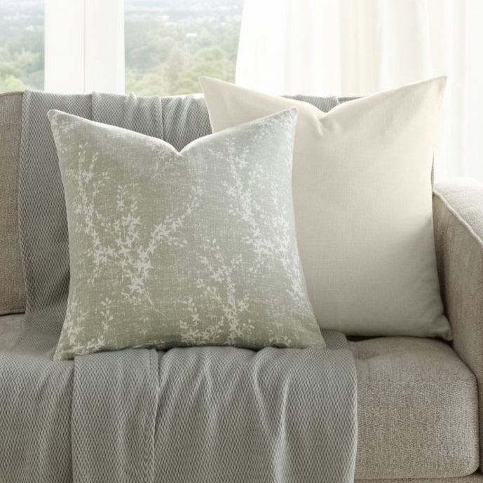 A neutral-toned sofa is adorned with two decorative pillows and a textured throw with fringe. A small wooden side table holds a vase with greenery. Large windows provide a scenic view in the background.