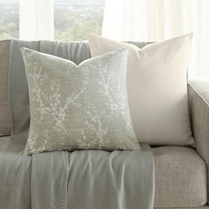A neutral-toned sofa is adorned with two decorative pillows and a textured throw with fringe. A small wooden side table holds a vase with greenery. Large windows provide a scenic view in the background.