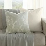A neutral-toned sofa is adorned with two decorative pillows and a textured throw with fringe. A small wooden side table holds a vase with greenery. Large windows provide a scenic view in the background.