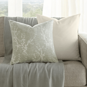 A neutral-toned sofa is adorned with two decorative pillows and a textured throw with fringe. A small wooden side table holds a vase with greenery. Large windows provide a scenic view in the background.
