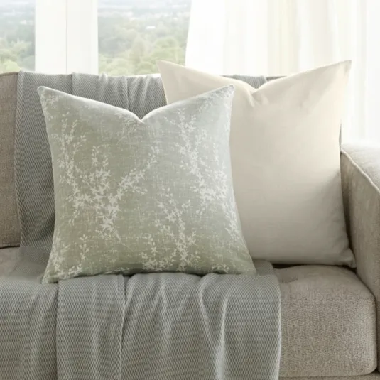 A neutral-toned sofa is adorned with two decorative pillows and a textured throw with fringe. A small wooden side table holds a vase with greenery. Large windows provide a scenic view in the background.
