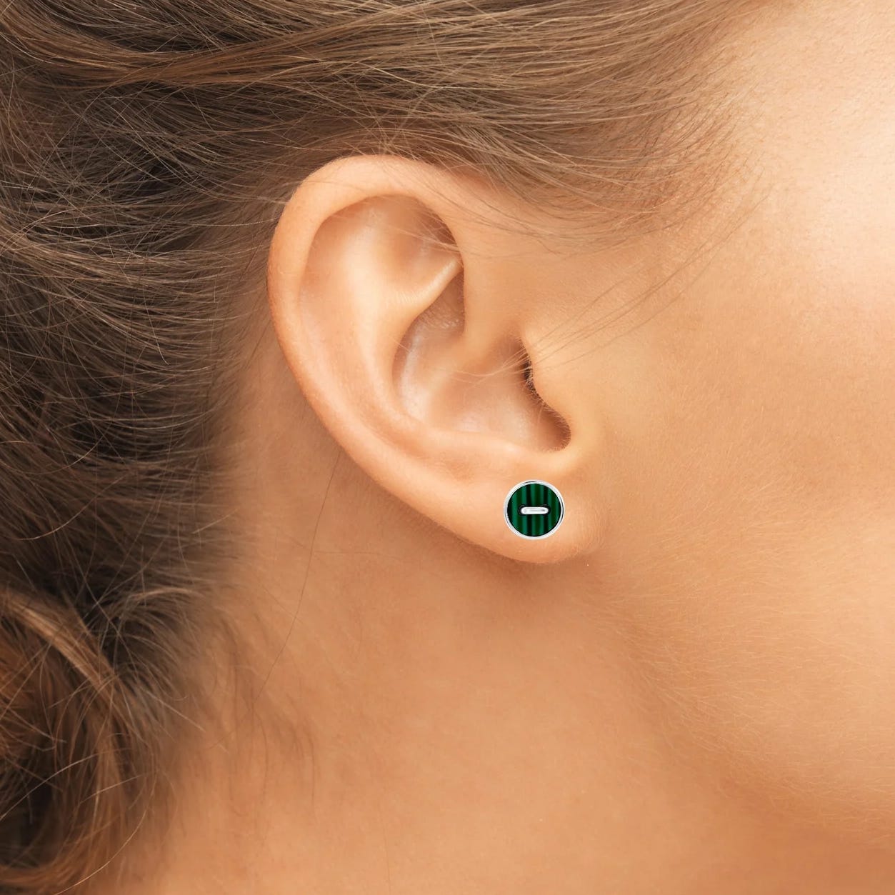 A close-up of a woman's ear wearing a small, round, green and silver stud earring. The earring features a minimalist, horizontal line design.
