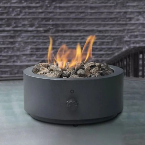 A round tabletop fire pit with lava rocks and an adjustable flame knob, set on a glass surface in an outdoor setting.