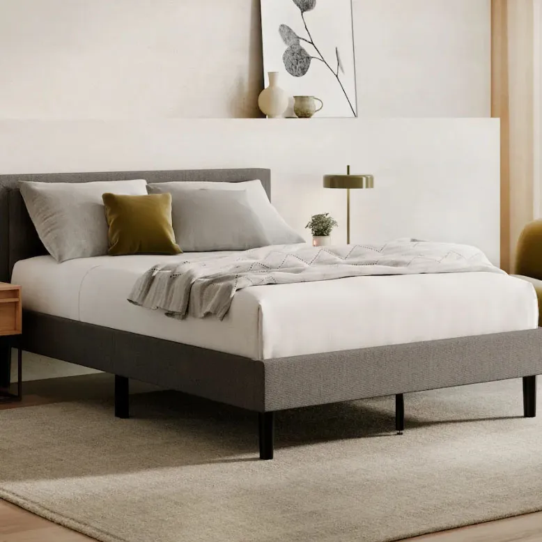 A neatly made bed with a gray upholstered frame and white bedding is shown in a modern room. It features gray and mustard pillows, a textured throw blanket, and is complemented by a nightstand, lamp, and minimalist decor.