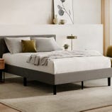 A neatly made bed with a gray upholstered frame and white bedding is shown in a modern room. It features gray and mustard pillows, a textured throw blanket, and is complemented by a nightstand, lamp, and minimalist decor.