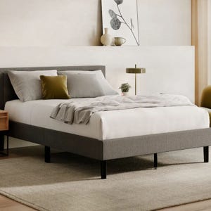A neatly made bed with a gray upholstered frame and white bedding is shown in a modern room. It features gray and mustard pillows, a textured throw blanket, and is complemented by a nightstand, lamp, and minimalist decor.