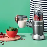 A NutriBullet blender with a silver and black base is blending blueberries and raspberries; to the left, a bowl of spinach and pomegranate seeds are visible.