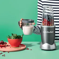 A NutriBullet blender with a silver and black base is blending blueberries and raspberries; to the left, a bowl of spinach and pomegranate seeds are visible.