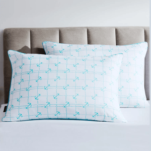 Two rectangular pillows with a repeating pattern of blue anchors are placed on a bed with a taupe upholstered headboard.
