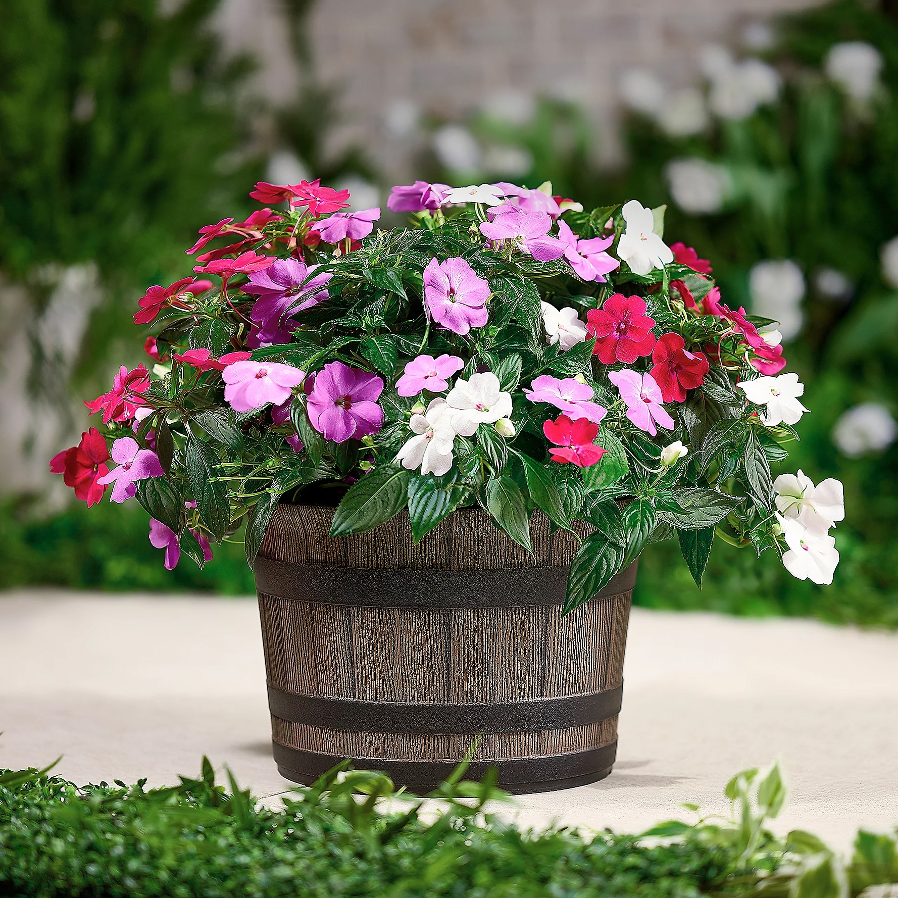A round wooden whiskey barrel planter contains a lush arrangement of vibrant flowers in shades of pink, purple, red, and white, with abundant green foliage, situated outdoors on a patio with greenery in the background.