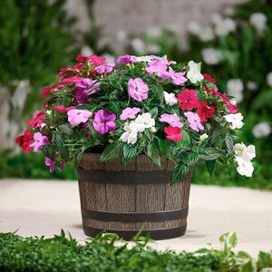 A round wooden whiskey barrel planter contains a lush arrangement of vibrant flowers in shades of pink, purple, red, and white, with abundant green foliage, situated outdoors on a patio with greenery in the background.