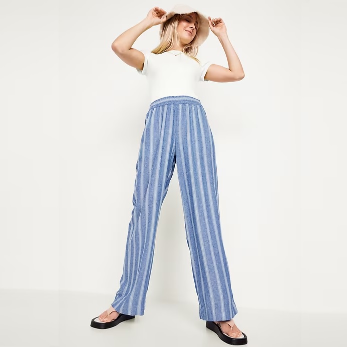 A person wearing a white sleeveless top and blue striped wide-leg trousers with black open-toe slides.